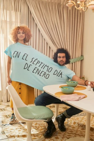 A man and woman sharing one oversized light blue t-shirt with Spanish text printed on the front, seated at a dining table in a retro room.