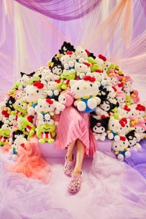 Person in a pink dress and Crocs sits in a pile of Hello Kitty and Sanrio character plushies against a pink and purple background.
