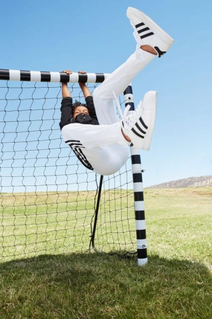 A young person in sportswear hangs upside down on a soccer goalpost outside on a sunny day, modeling a sporty, playful, active lifestyle.