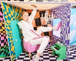 Woman in white hoodie and sweatpants posing inside a surreal miniature dollhouse set designed by Todd Davis