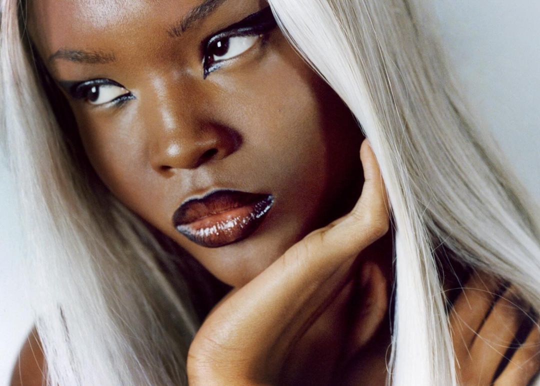 Close-up portrait of a young Black woman with silver hair, wearing glossy lipstick and black eyeliner, with her hand near her face.