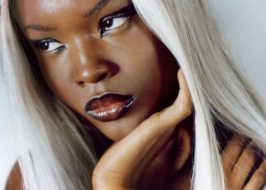 Close-up portrait of a young Black woman with silver hair, wearing glossy lipstick and black eyeliner, with her hand near her face.