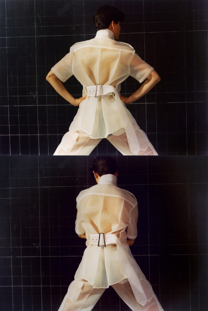 Two images of a person in a white, translucent short-sleeved shirt, belt and pants, standing with hands on hips against a black tile wall.