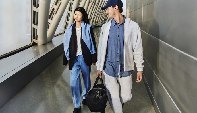 Two people walk in a modern, brightly lit indoor space. The woman is in blue pants, a black blazer, and the man wears a baseball cap.