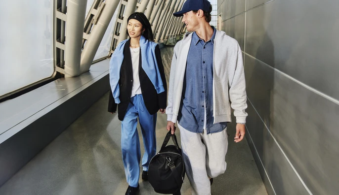 Two people walk in a modern, brightly lit indoor space. The woman is in blue pants, a black blazer, and the man wears a baseball cap.