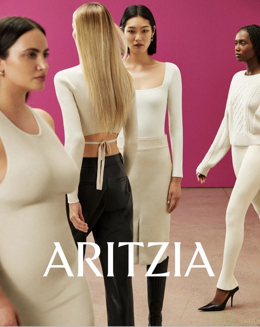 Four female fashion models showcase neutral-toned Aritzia clothing in a studio setting against a pink backdrop, including dresses and pants.
