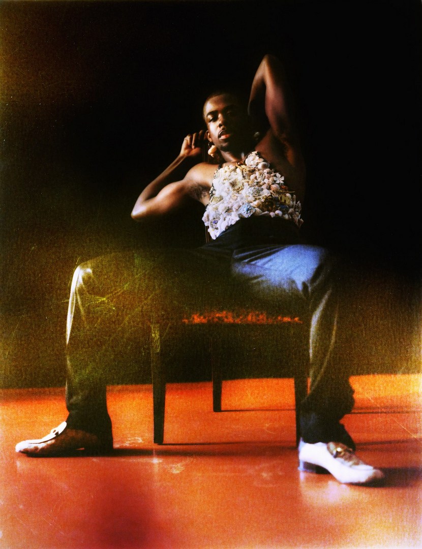 Elegant pose: muscular African-American man sits on chair in dark room, wearing jeans, white shoes, and decorative top.