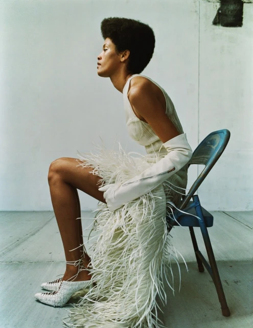 Side profile view of a fashionable Black female model in an ivory dress with feathers, gloves, and heels sitting on a blue chair.