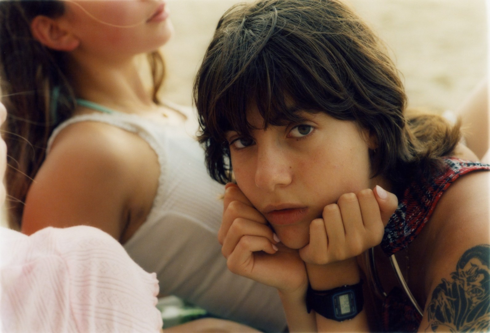 Close-up portrait of a young woman resting her chin on her hands outdoors with another person partially visible in the background