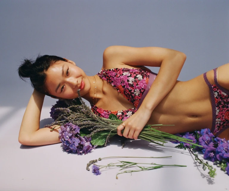 A young Asian woman lies on a white surface in a floral bikini, gazing at the camera and holding a bouquet of purple flowers.