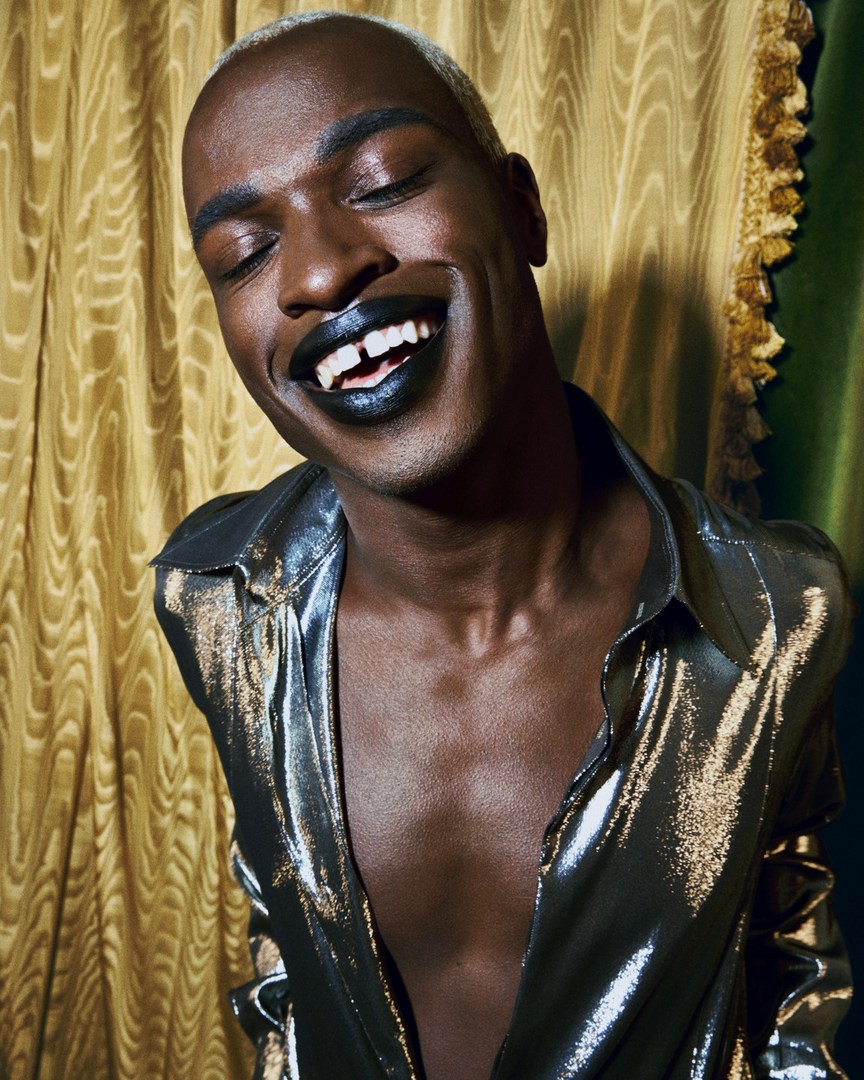 Close-up studio portrait of an adult man with short bleached hair wearing a metallic silver shirt and dark lipstick, against a textured backdrop.