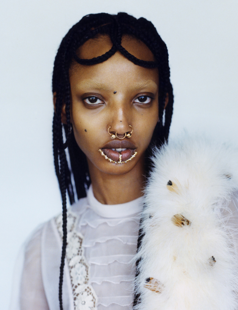 Close-up of a woman with braided hair and nose rings, wearing a white lace top and feathered boa against a bright, neutral background.