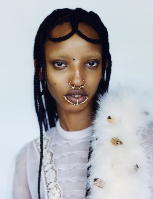 Close-up of a woman with braided hair and nose rings, wearing a white lace top and feathered boa against a bright, neutral background.