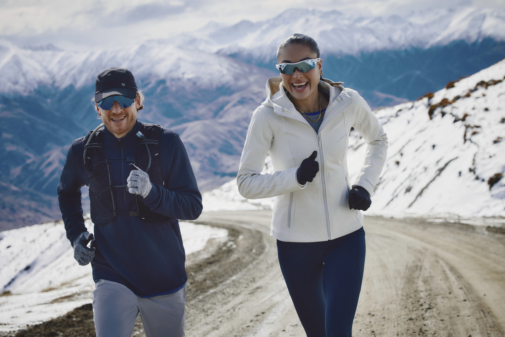 Two people run on a mountain road. The man wears a black cap and jacket, the woman a white jacket and sunglasses, snowy mountains behind.