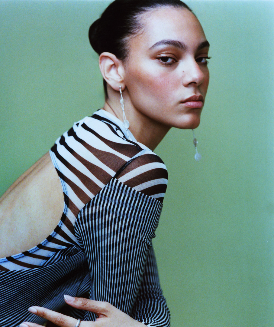 Portrait of a slender adult woman in a backless black and white striped top against a solid green background in a studio setting.