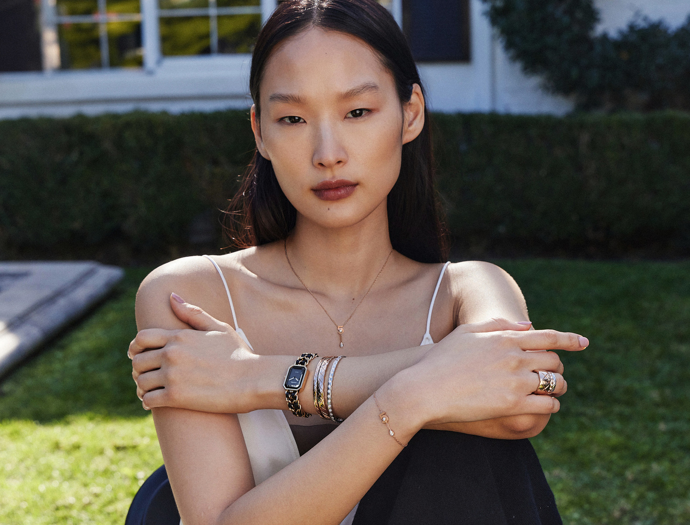 Asian woman in a tank top poses outdoors with jewelry, including a watch and bracelets, against a green lawn and white house.
