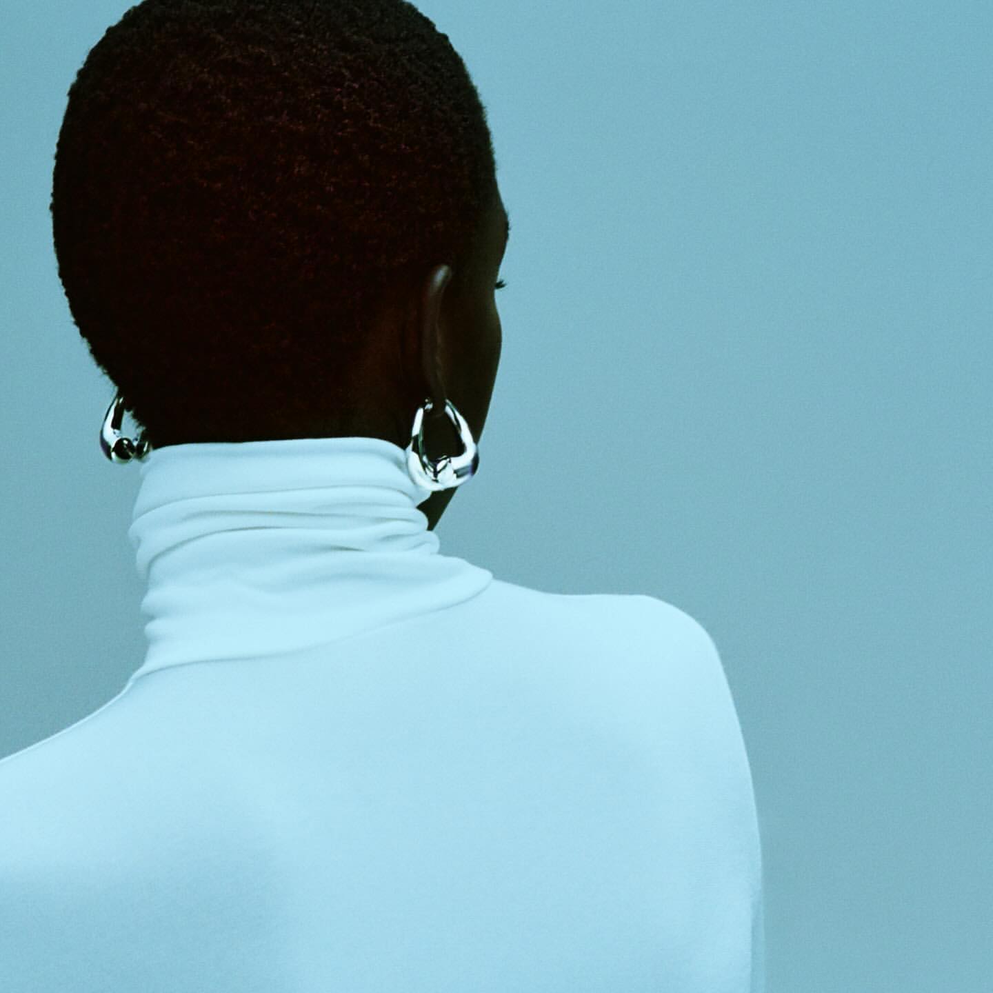 Rear view of a person with short dark hair wearing a white turtleneck and chunky silver earrings against a blue background.