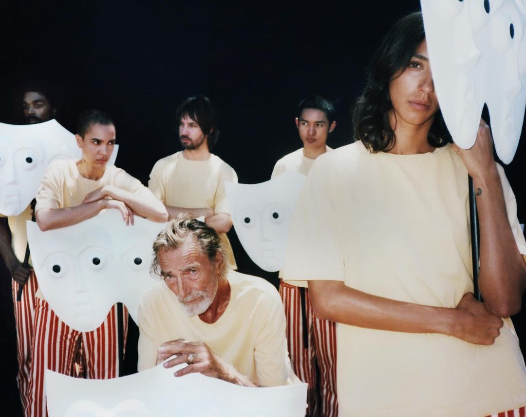 Group of people in yellow shirts and striped pants holding large, white, mask-like props against a dark background, artistic and mysterious.