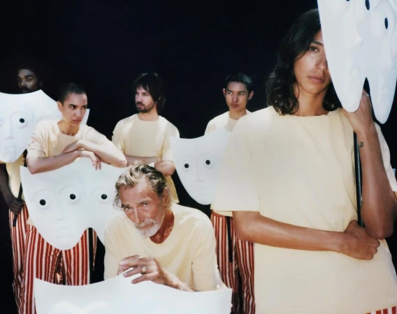 Group of people in yellow shirts and striped pants holding large, white, mask-like props against a dark background, artistic and mysterious.