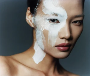 Close-up portrait of a woman with bold white paint brushed across the right side of her face and neck, photographed by Sam Nixon.