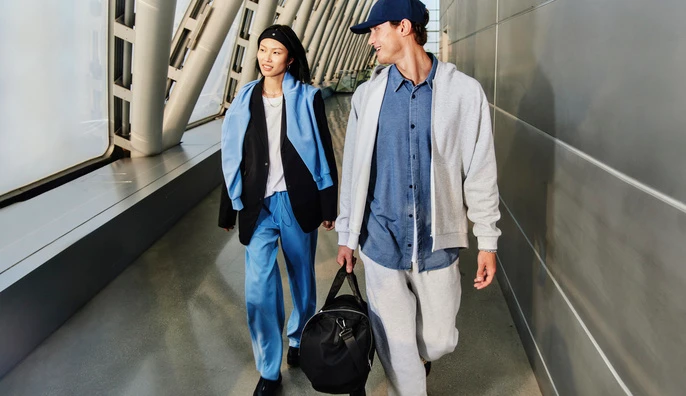 Two models walking together through a modern transit terminal, carrying a black duffel bag and wearing casual clothing.