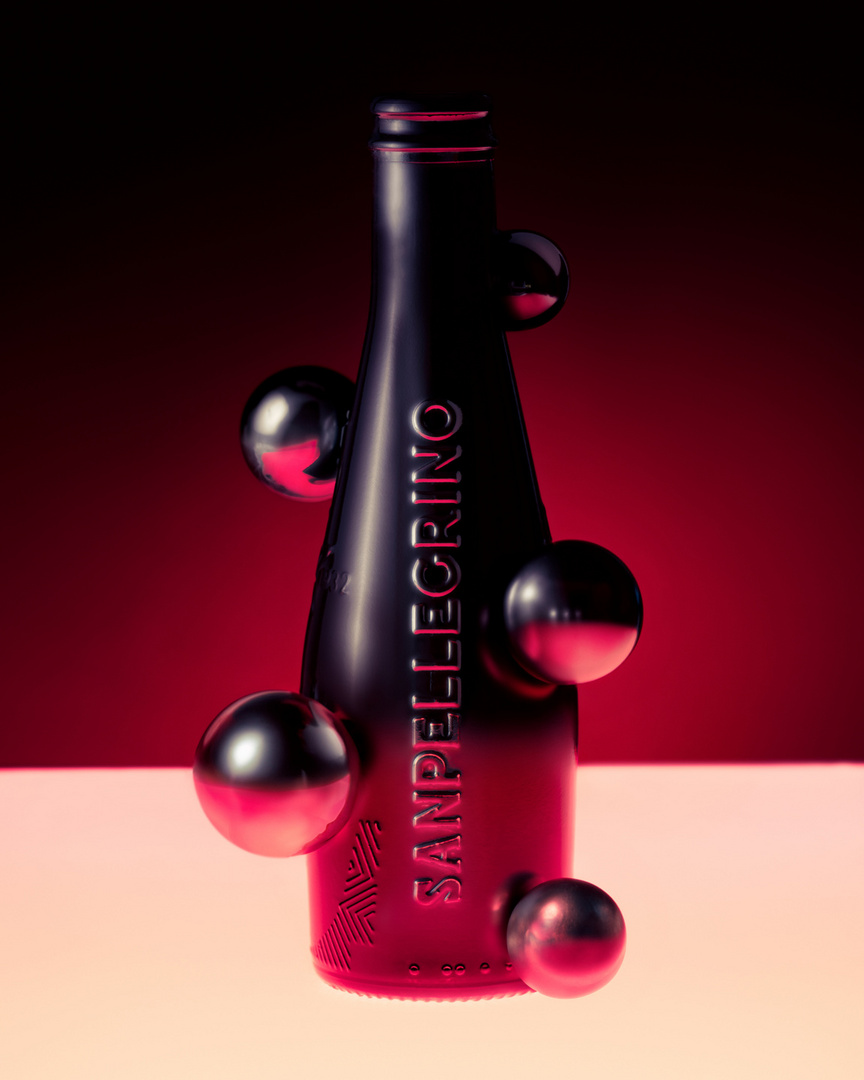 Elegant dark glass San Pellegrino bottle surrounded by metallic spheres, set against a gradient red and white backdrop.