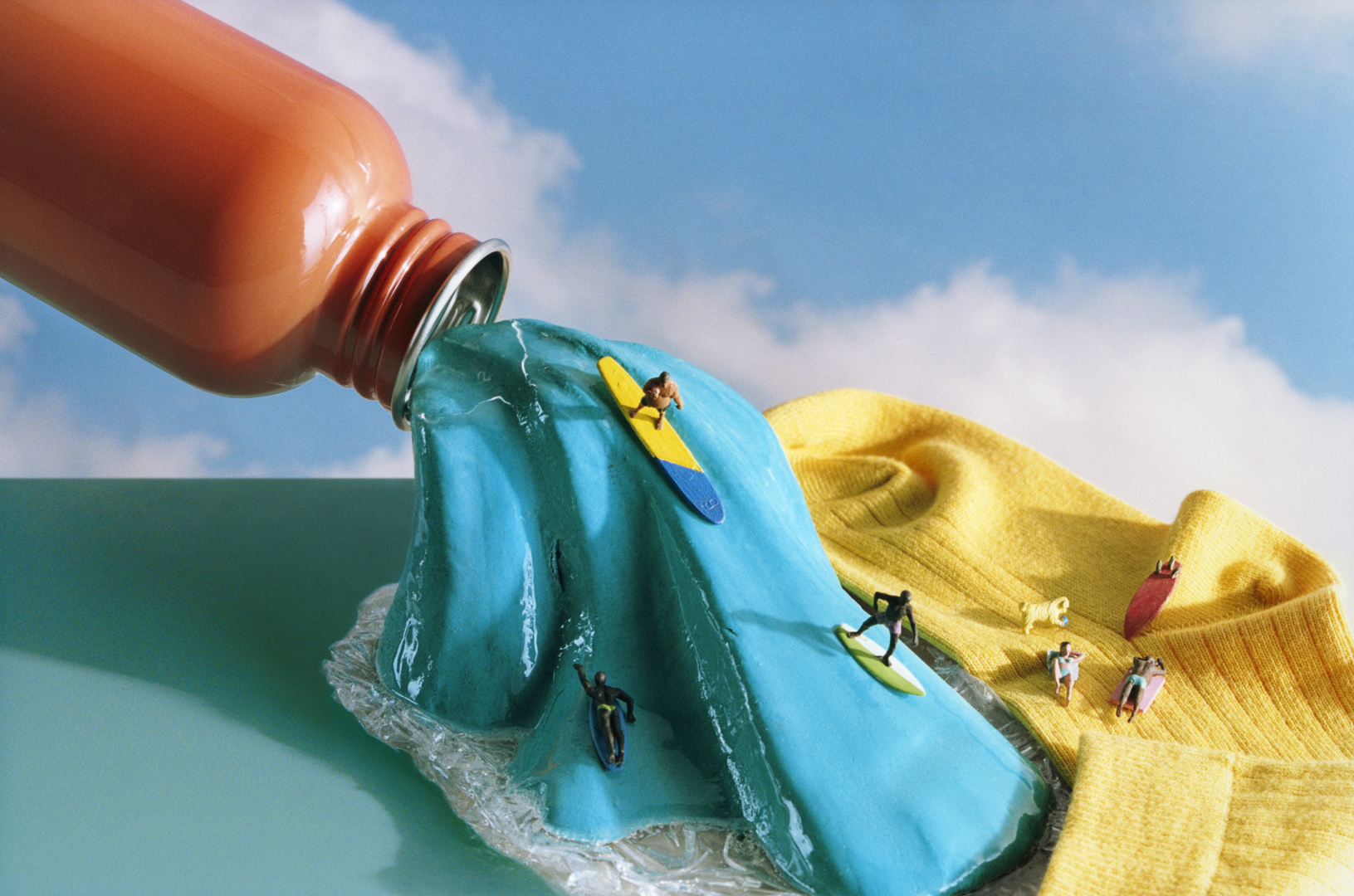 Miniature surfers ride a teal liquid wave pouring from an oversized orange bottle, beside a yellow sock and under a cloudy blue sky.