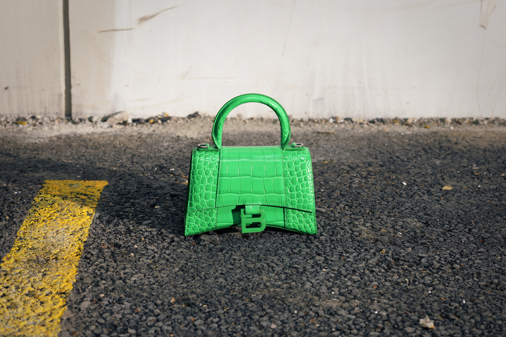 Bright green Balenciaga handbag with crocodile texture, situated on an asphalt surface with a yellow painted line, next to a white wall.