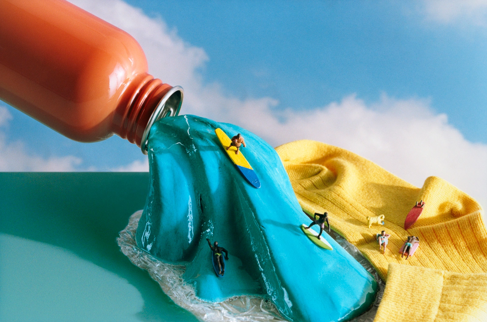 Miniature surfers ride a glossy blue resin wave pouring from an orange bottle next to a yellow fabric surface.
