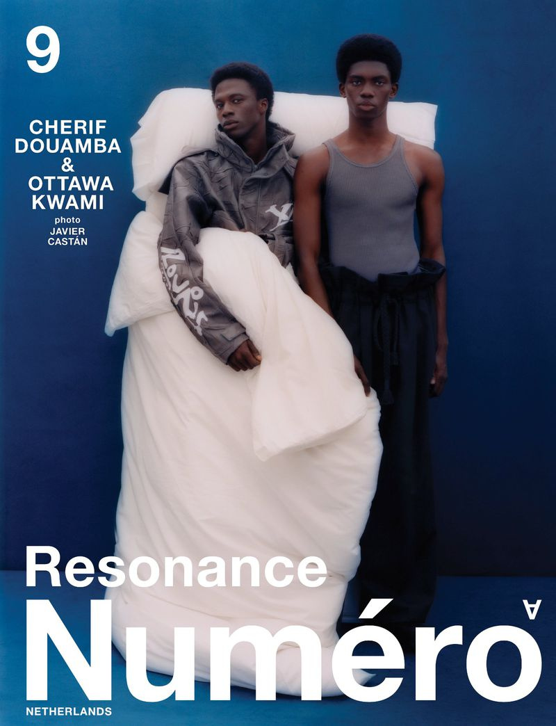 Two black men posing against a solid blue background, with one draped in a large white duvet and wearing a patterned jacket.