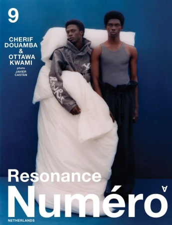 Two black men posing against a solid blue background, with one draped in a large white duvet and wearing a patterned jacket.
