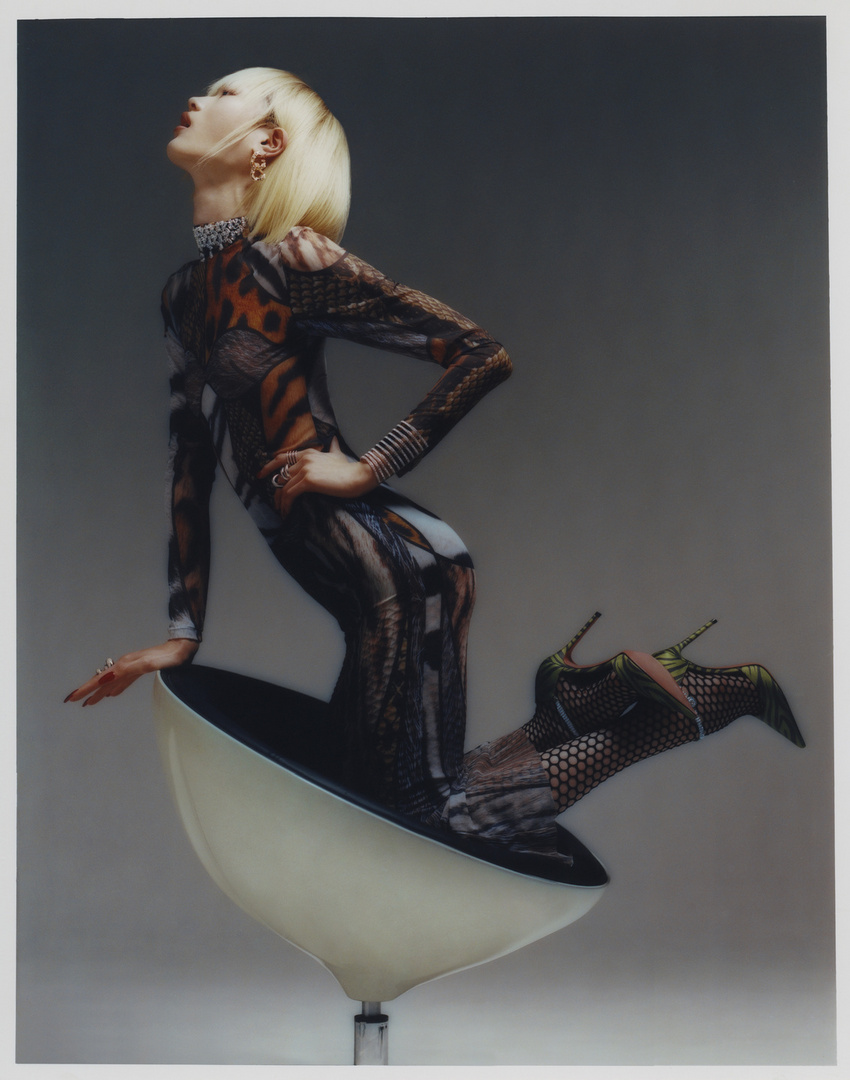 Model with blonde hair wearing patterned jumpsuit and heels kneeling inside a modern bowl chair