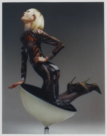 Model with blonde hair wearing patterned jumpsuit and heels kneeling inside a modern bowl chair