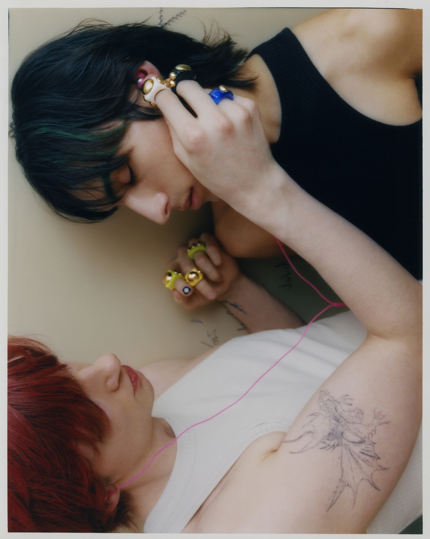 Two people in tank tops and rings pose against a cream wall, one lying down and the other leaning over, with pink string in the shot.