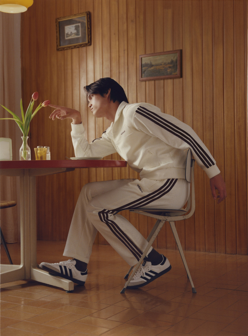 Man wearing a white track jacket and matching track pants seated at a wooden table, gently touching flowers in a vase.