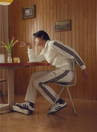 Man wearing a white track jacket and matching track pants seated at a wooden table, gently touching flowers in a vase.