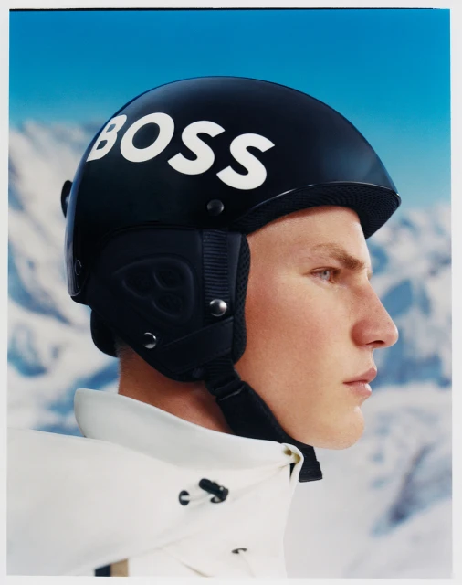 Profile shot of a male model wearing a black BOSS-branded ski helmet and white jacket against a blurred mountain and sky background.