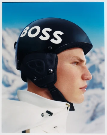 Profile shot of a male model wearing a black BOSS-branded ski helmet and white jacket against a blurred mountain and sky background.
