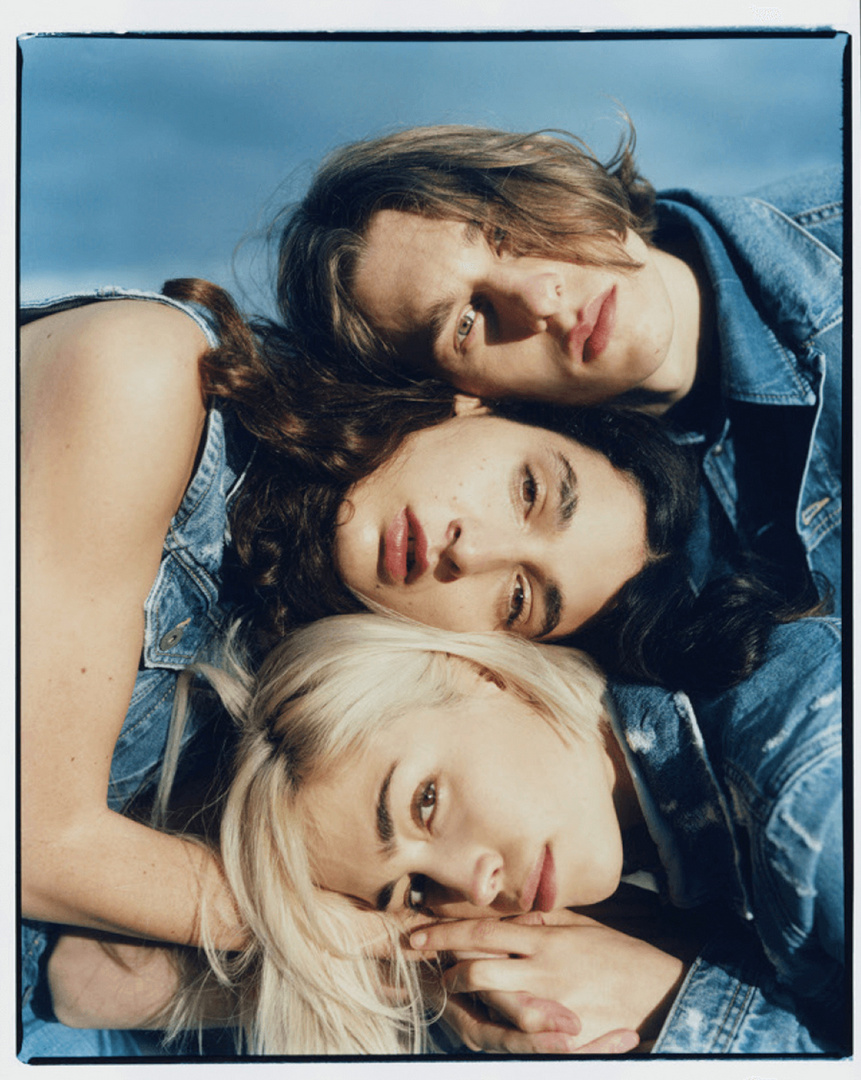 Three fair-skinned women in denim jackets pose outdoors against a blue sky, creating a fashion-forward, youthful vibe.