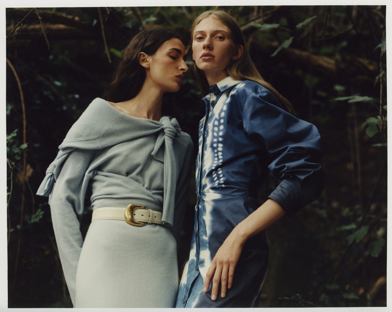 Two women in stylish outfits pose in a lush forest. One wears a light blue dress with a knotted shawl and belt, the other a blue tie-dye dress.