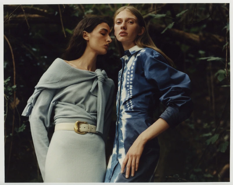 Two women in stylish outfits pose in a lush forest. One wears a light blue dress with a knotted shawl and belt, the other a blue tie-dye dress.