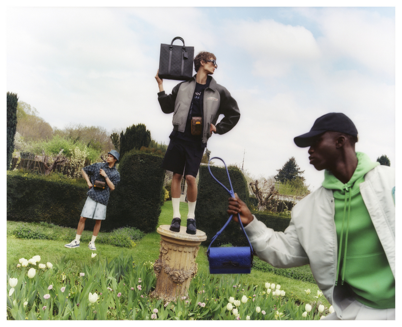 Three men model handbags and accessories in an outdoor garden setting with trees, lawn, and flowers under a bright daytime sky.