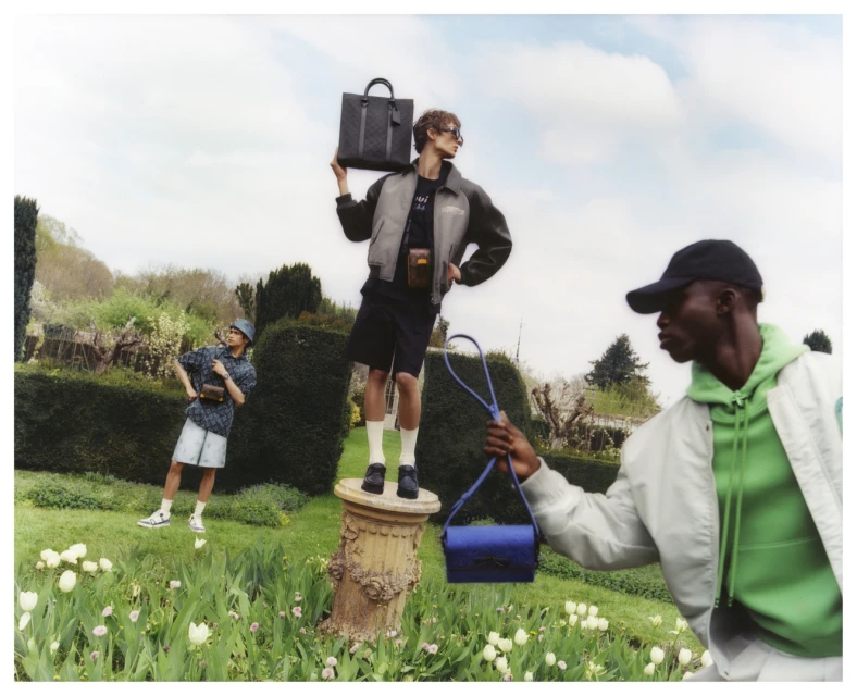Three men model handbags and accessories in an outdoor garden setting with trees, lawn, and flowers under a bright daytime sky.