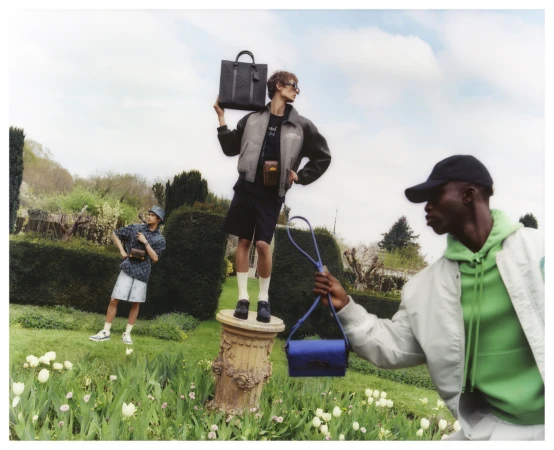 Three men model handbags and accessories in an outdoor garden setting with trees, lawn, and flowers under a bright daytime sky.