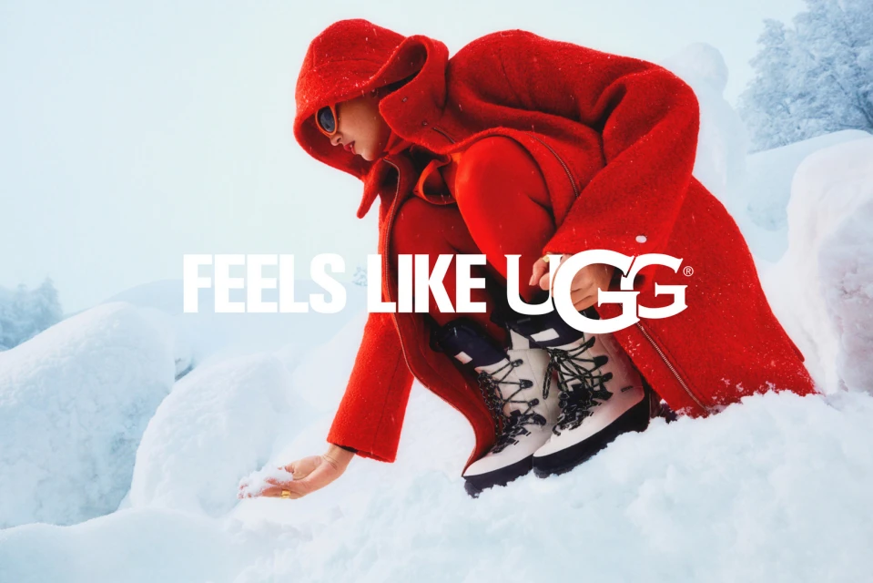 Woman in red hooded coat squatting on snow, touching snow with gloved hand, wearing UGG boots in a snowy landscape.