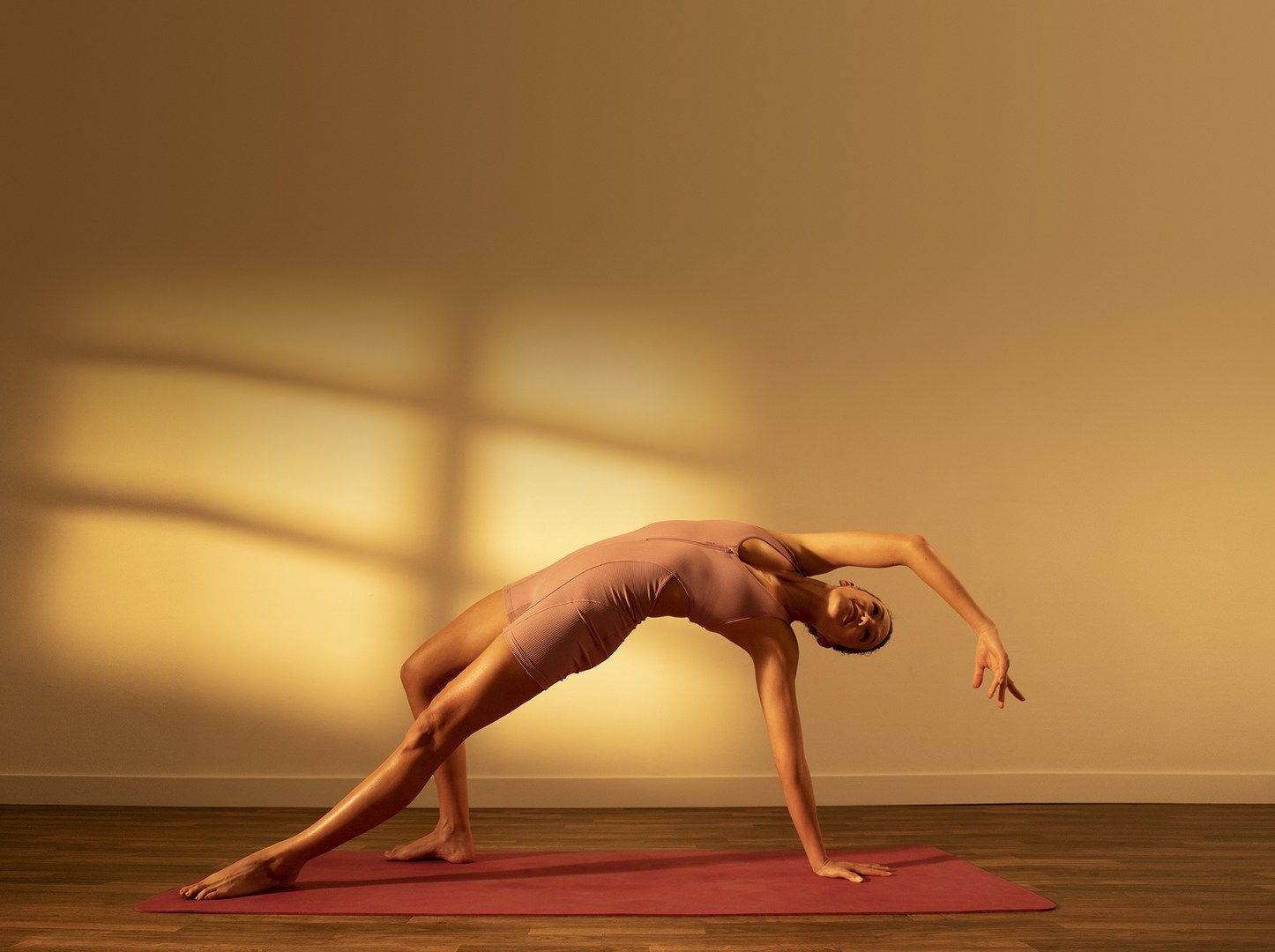 Adult woman in a pink bodysuit doing a yoga pose on a red mat indoors with warm lighting and a window shadow on the wall.