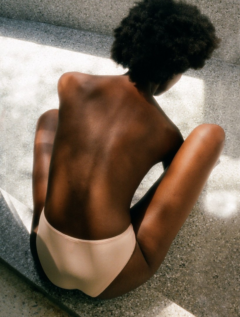 Back view of a slim, tan-skinned woman with an Afro hairstyle, sitting on a concrete surface in sunlight, wearing neutral-colored underwear.