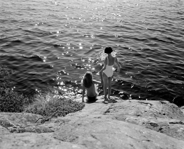 Two females in swimsuits stand on rocks looking at the ocean in a black and white photograph with sparkling water and rocky outcrops.