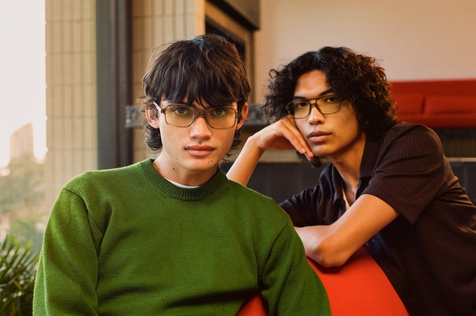 Two young adult males wearing glasses, one in a green sweater, the other in brown, seated indoors near a window with natural light.