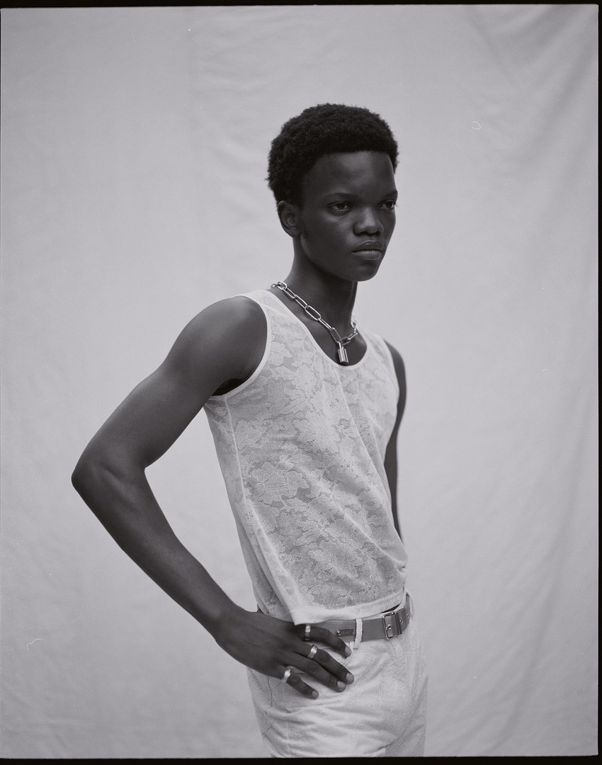 Portrait of a young adult Black male in a white tank top and light-colored pants, posing against a neutral background in a studio setting.